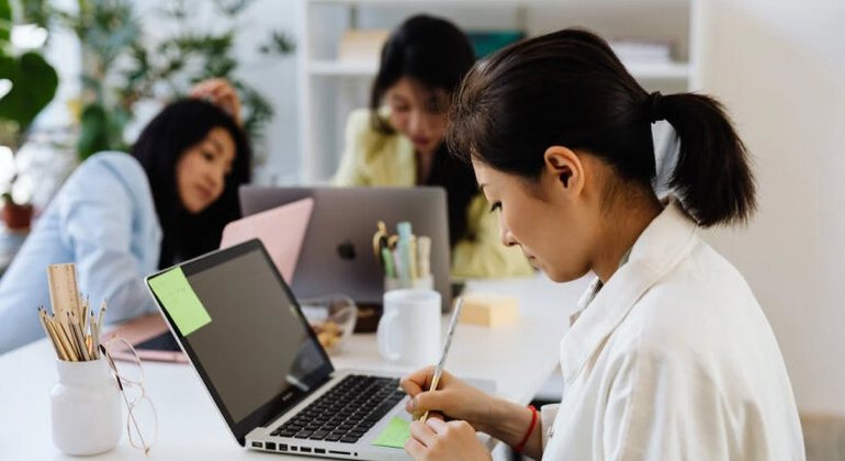 Professional team member organizing task notes on a laptop with colleagues nearby.